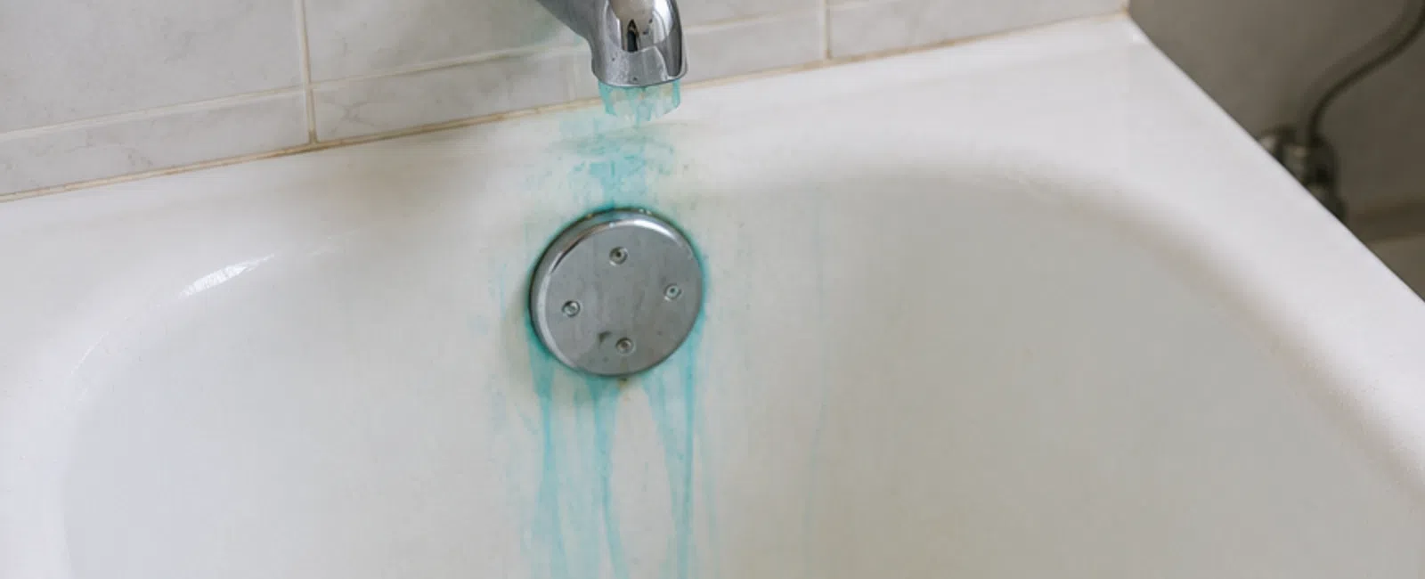 Blue-green stains from running water visible on bathtub, bathroom sink, and toilet bowl in a white tiled bathroom.