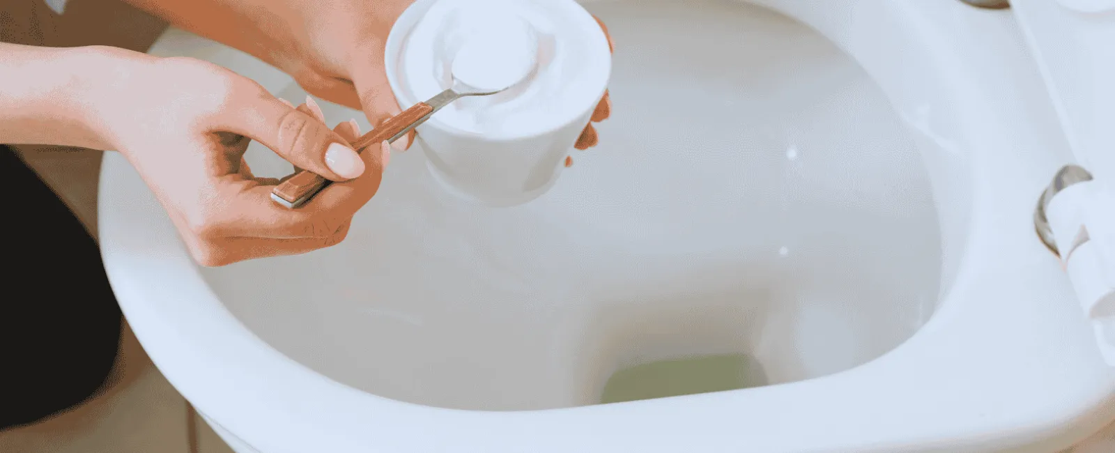 Person holding baking soda cup and spoon over a clean white toilet bowl for cleaning.