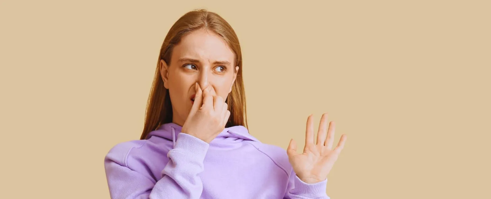 Young woman in purple hoodie holding nose and showing disgust on beige background