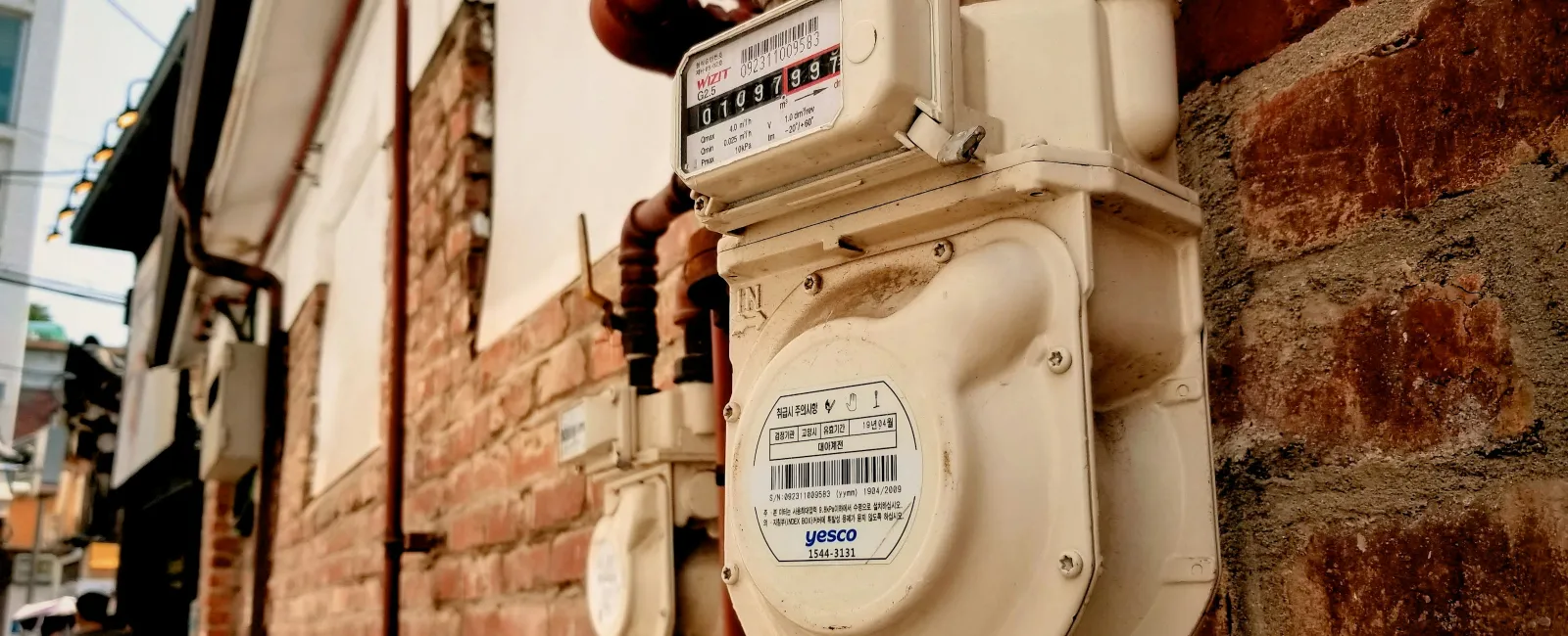Close-up of a white gas meter mounted on a brick wall with metal pipes and urban background.