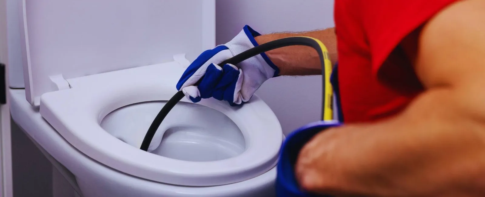 Plumber using a drain snake tool to unclog a white toilet wearing gloves and red shirt.
