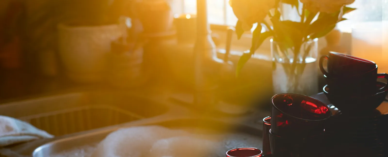 Sunlit kitchen sink filled with soapy water next to stacked red glass cups and a vase of white flowers on the counter
