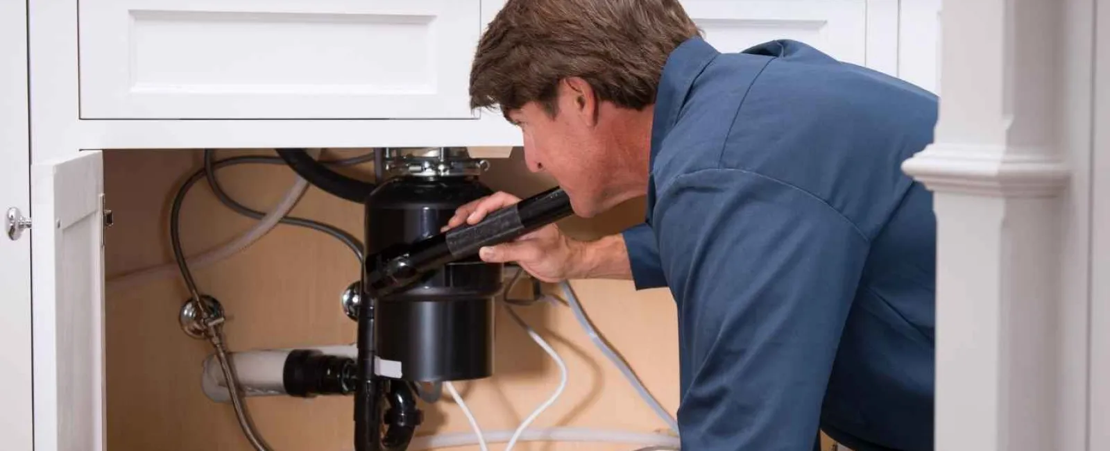 Technician in blue uniform inspecting kitchen sink plumbing and garbage disposal under white cabinet.
