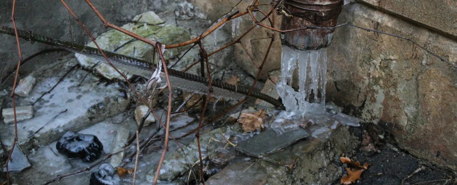 Rusty pipe with frozen water and icicles against aged concrete wall outdoors in winter.