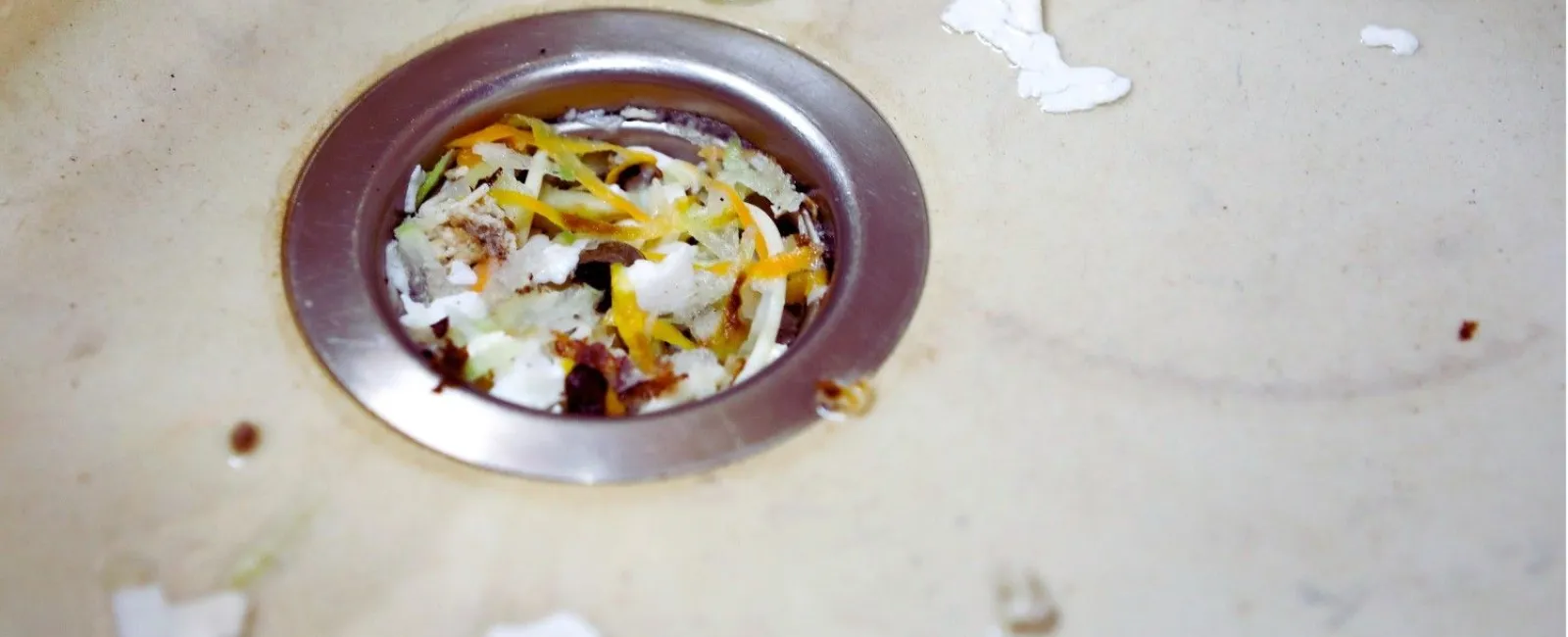 Close-up of a kitchen sink drain clogged with food scraps and vegetable peels on a beige surface.