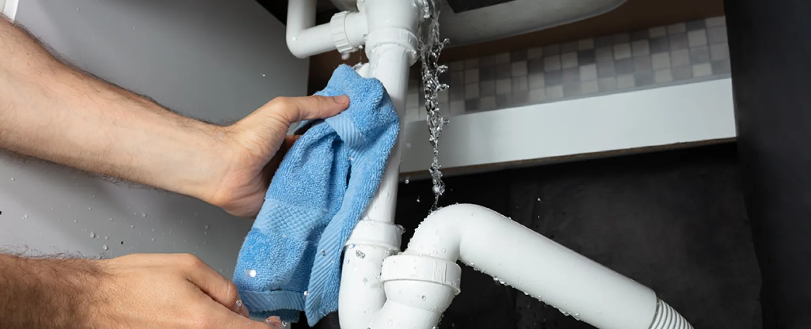 A person cleaning a leaking pipe under a kitchen sink with a blue cloth, water splashing.