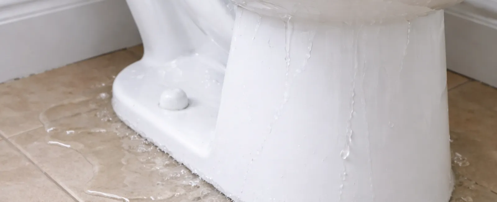 White toilet leaking water onto beige tiled bathroom floor causing a plumbing issue.