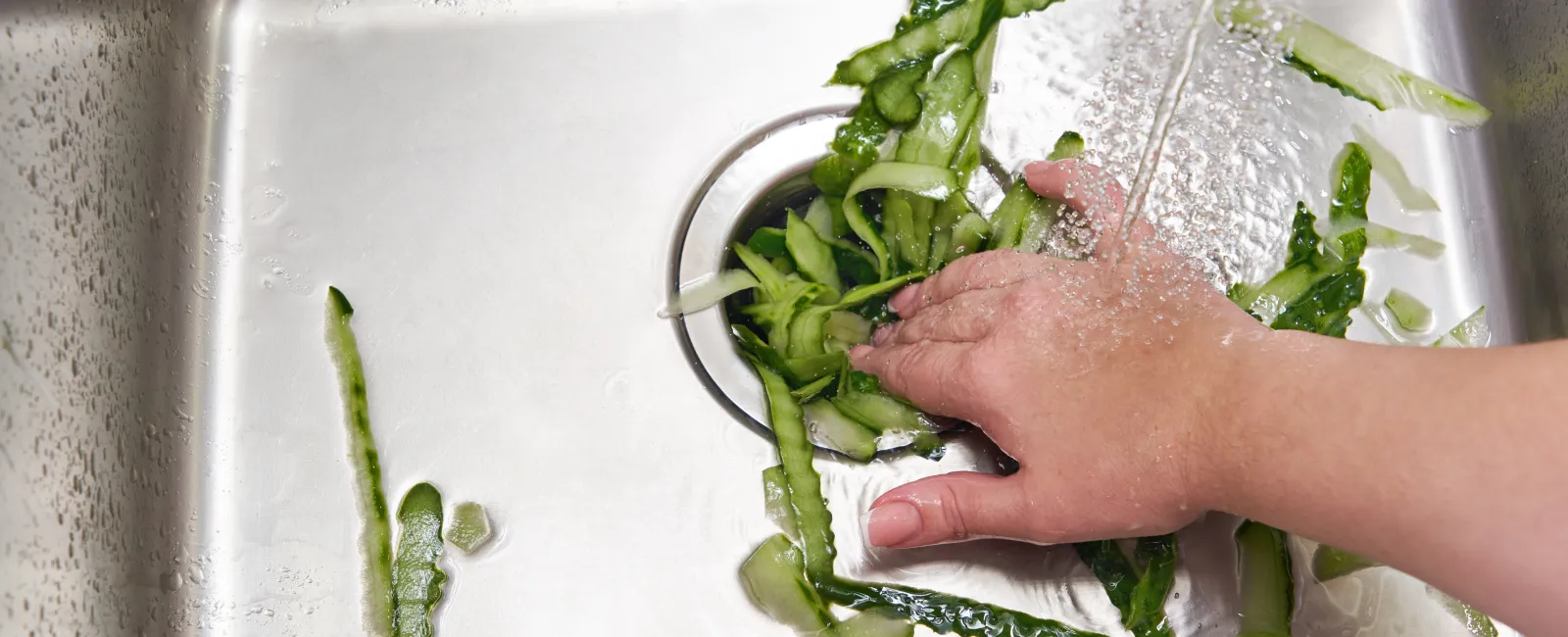 Hand washing green vegetable peels under running water in a stainless steel kitchen sink drain.