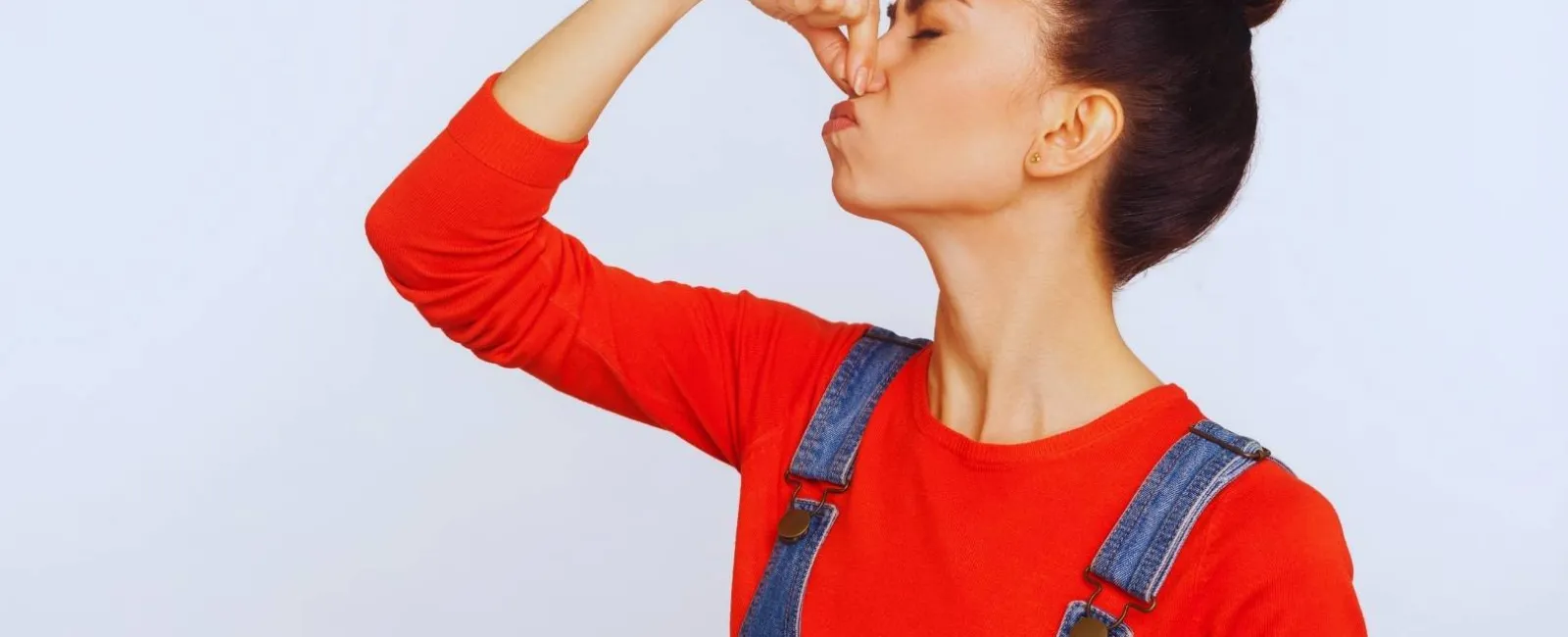 Young woman in red shirt and denim overalls pinching nose to block bad smell against a plain background
