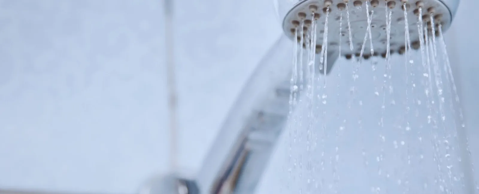 Close-up of a chrome showerhead with water droplets flowing against a tiled bathroom wall.