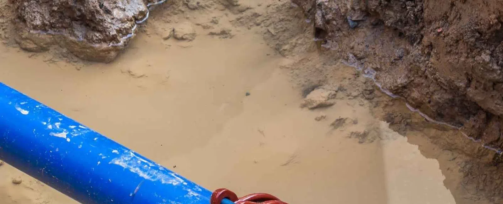 Blue and black water pipes connected by a red coupling in muddy trench with wet soil surrounding.