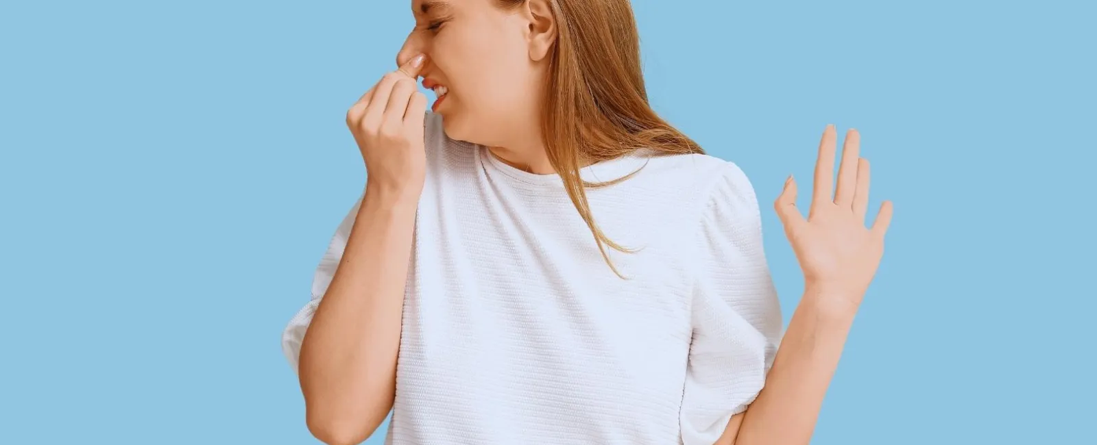 Young woman in white shirt holding her nose with a disgusted expression on a blue background