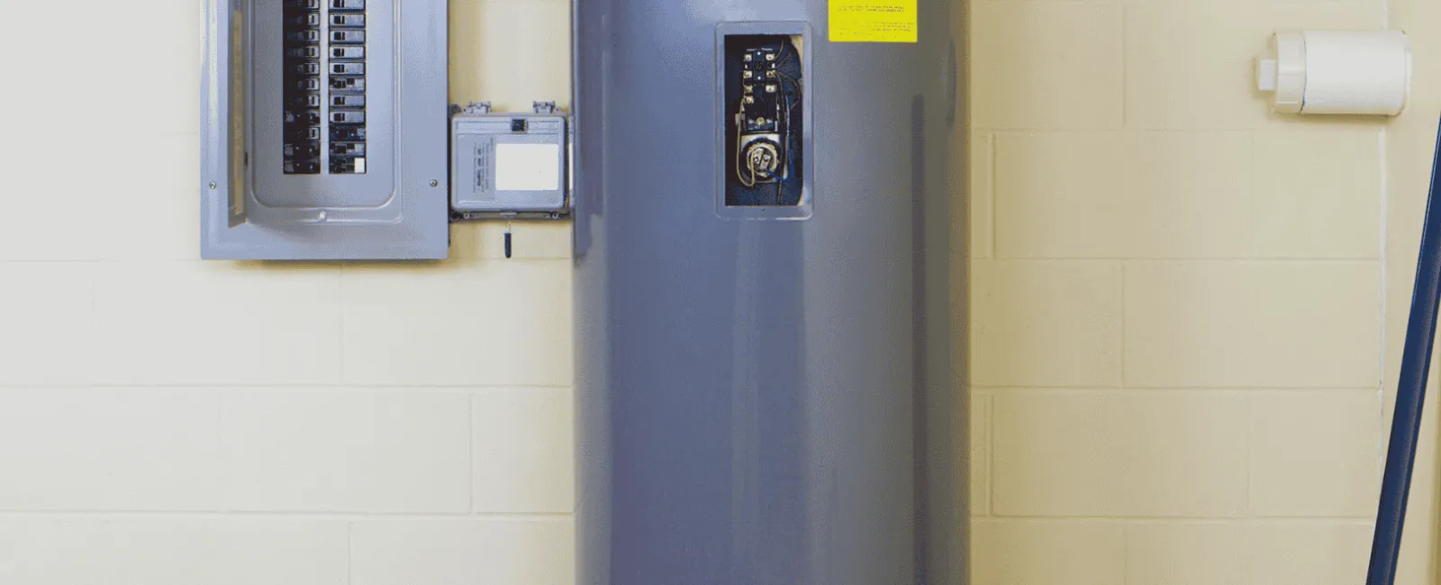 Large gray electric water heater next to a circuit breaker panel mounted on a beige wall in a utility room.