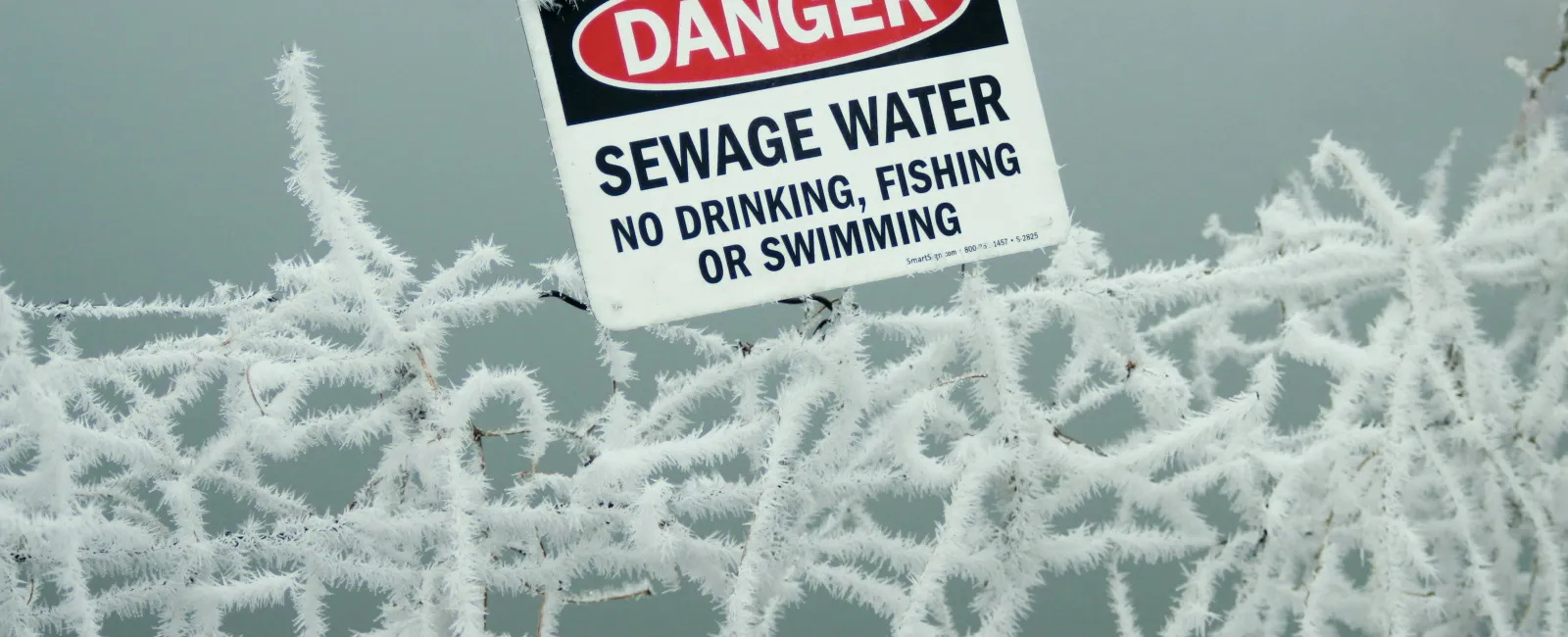 Danger sign warning of sewage water, no drinking, fishing, or swimming, on frost-covered wire fence.