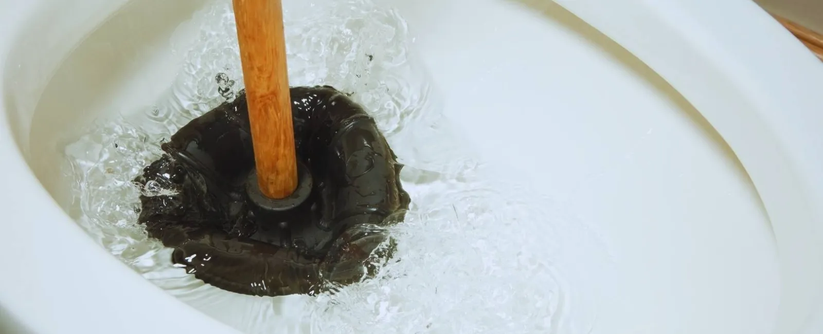 Close-up of a toilet bowl being unclogged with a wooden-handled plunger creating water pressure.