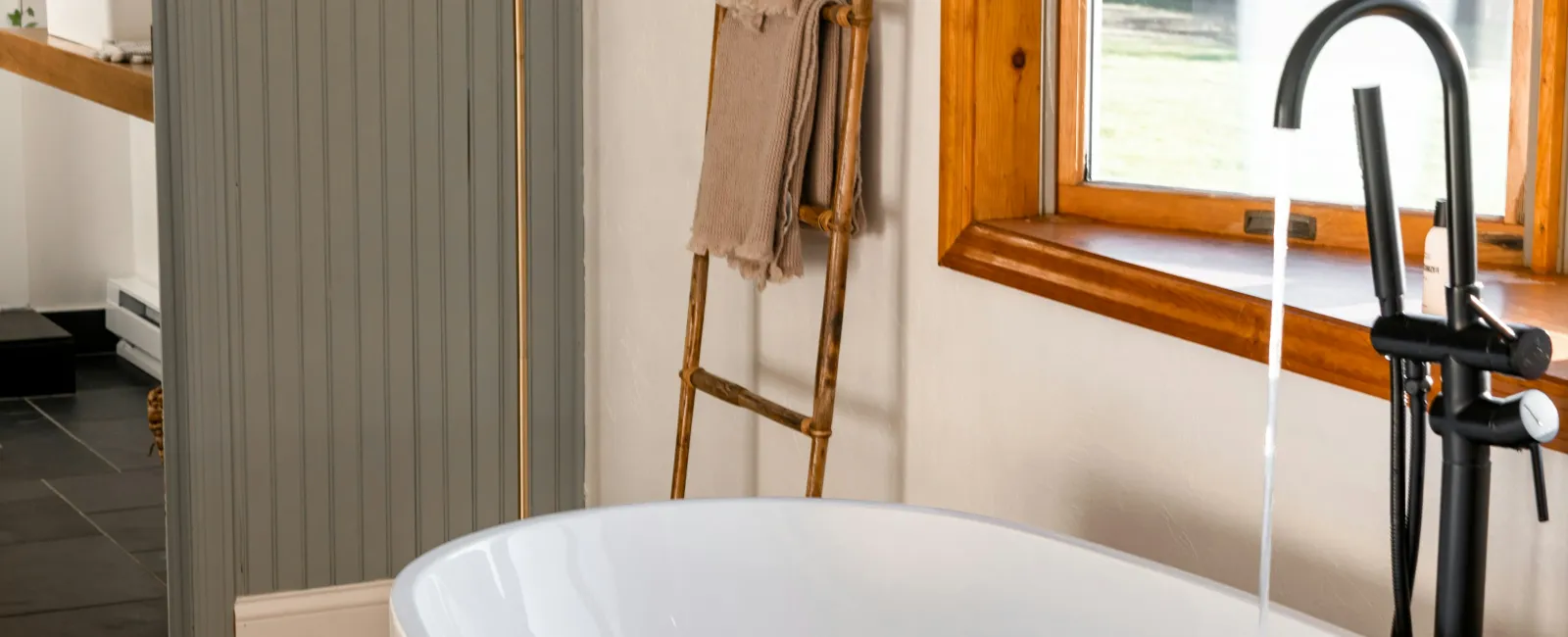 Modern freestanding white bathtub with running faucet, wooden ladder towel rack, and large window with natural light.