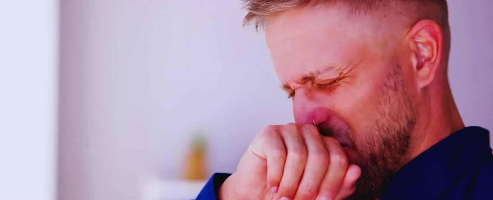 Man in blue shirt covering mouth and nose, showing signs of sneezing or illness indoors.