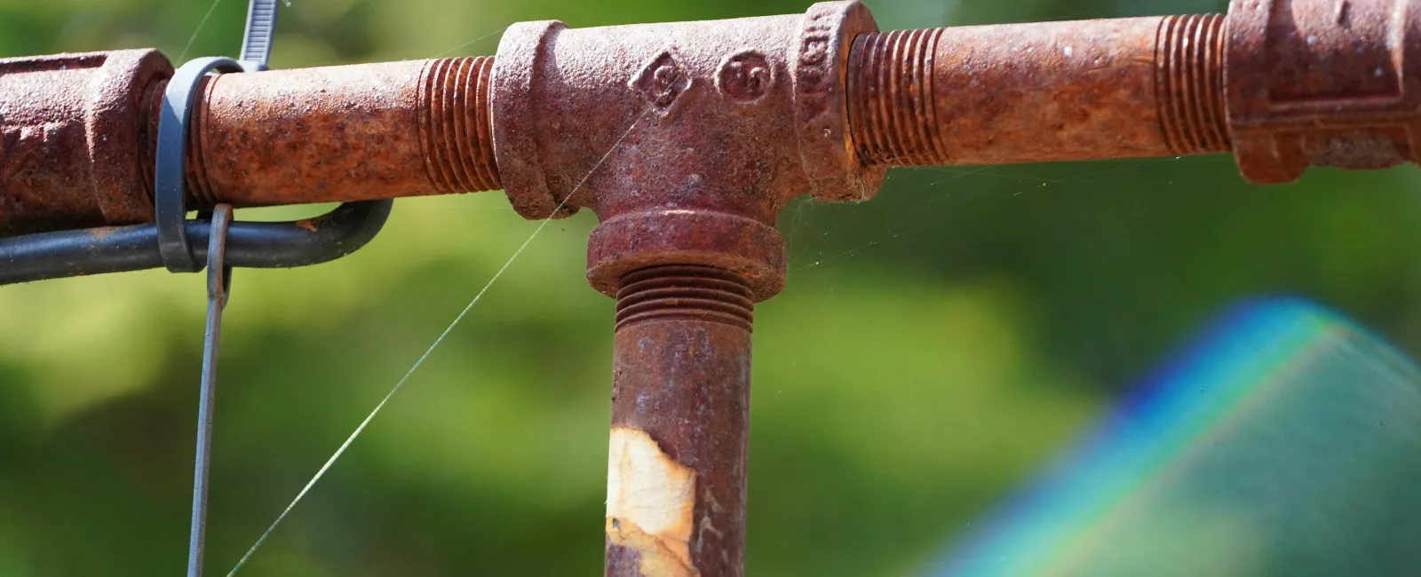 Close-up of a rusted metal pipe joint with zip tie and green blurred background.