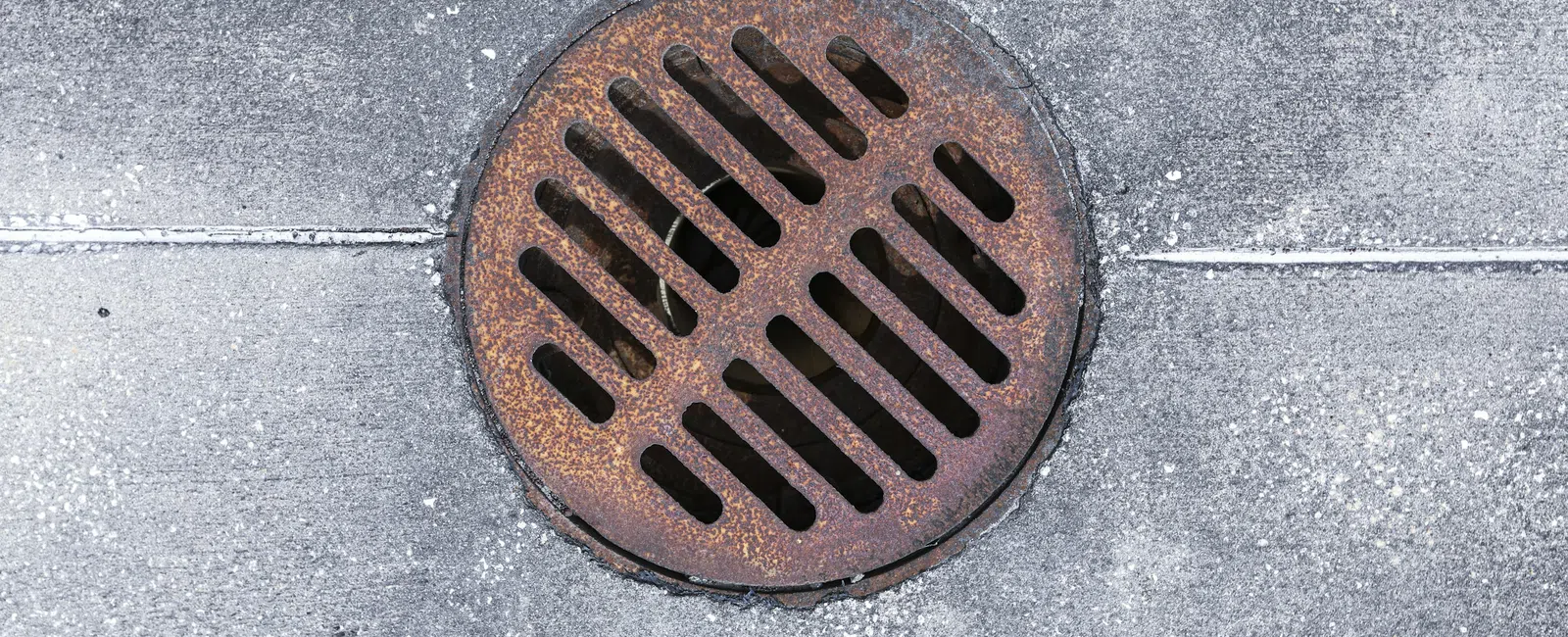 Rusty round metal drain cover embedded in gray concrete pavement with diagonal slits