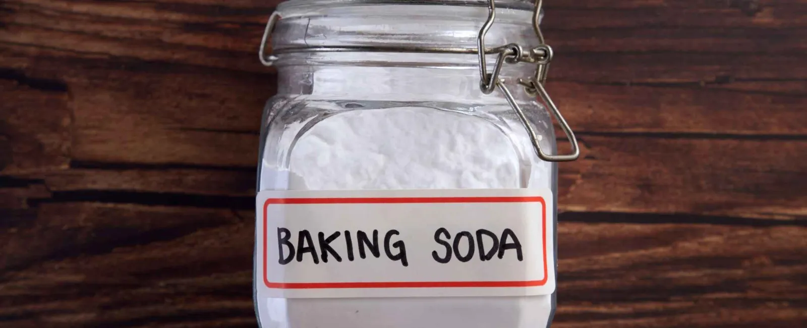 Glass jar filled with white powder labeled baking soda on a wooden surface background