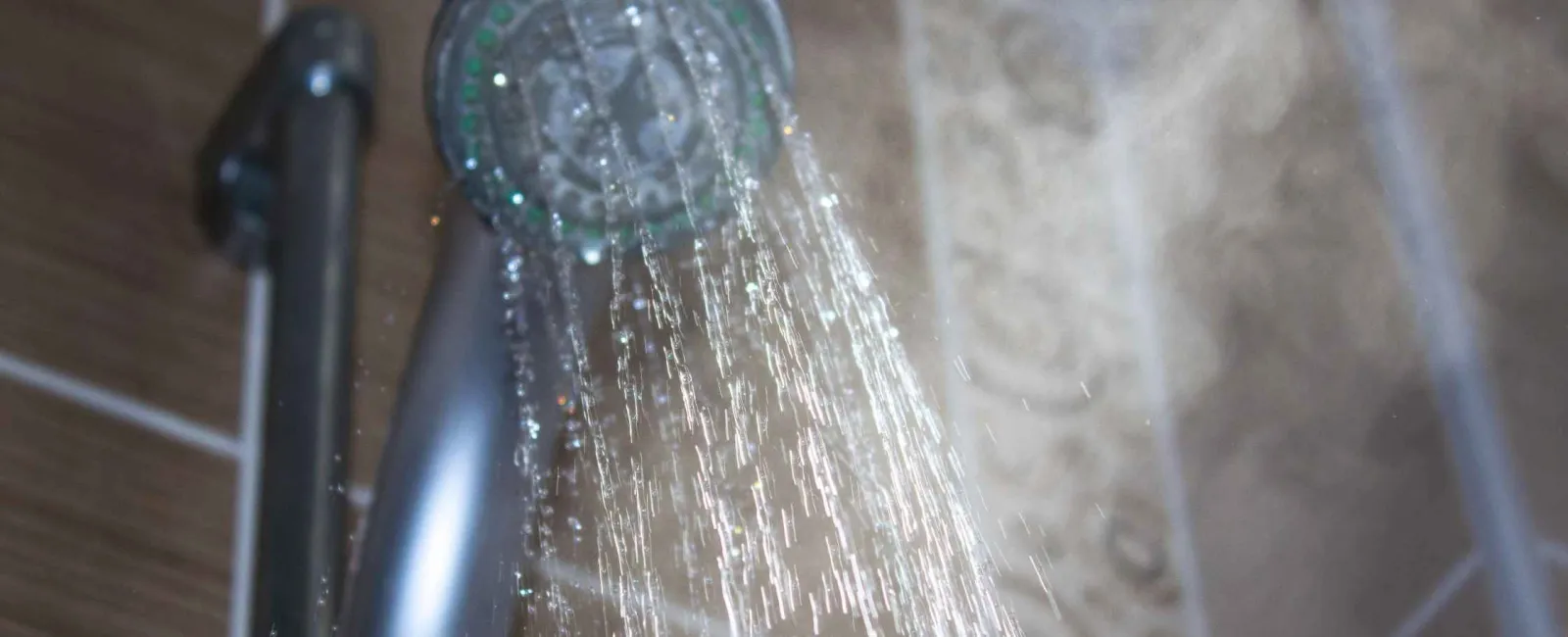 Close-up of water streaming from a silver showerhead with steam rising in a tiled bathroom.