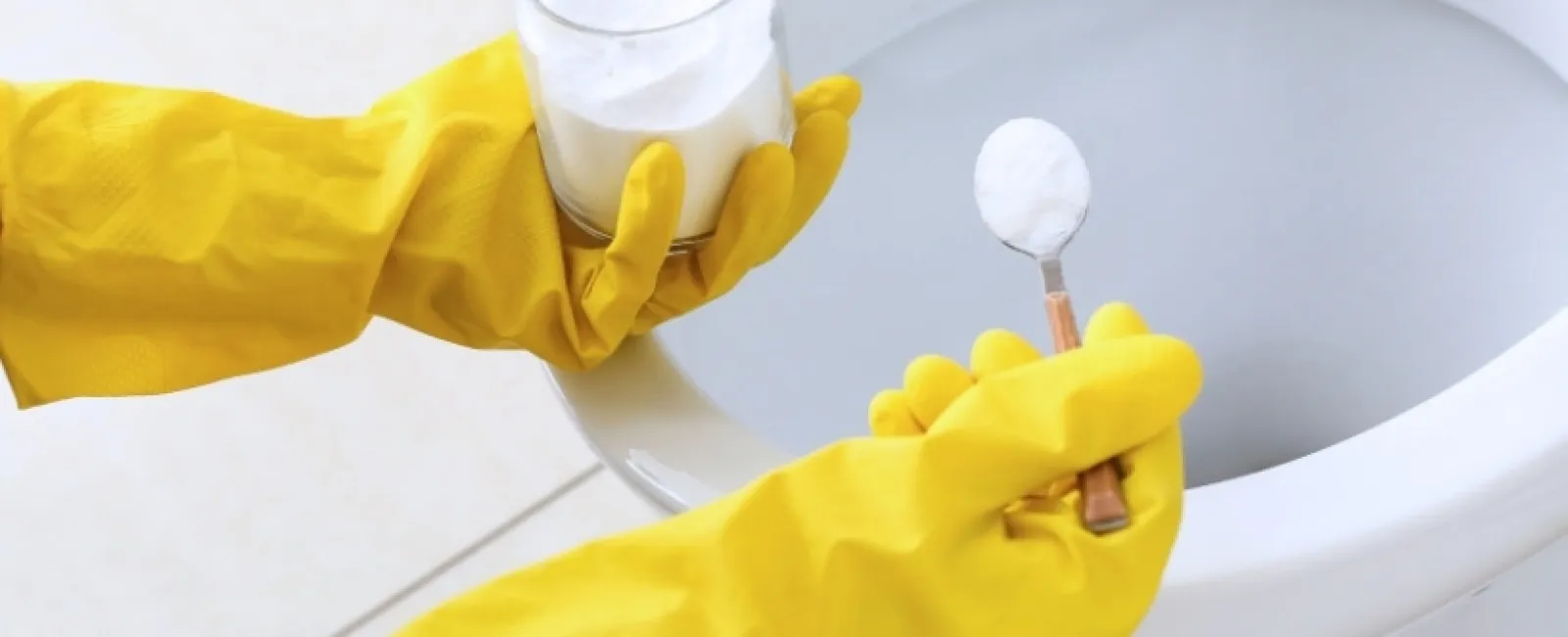 Person wearing yellow gloves holding baking soda to clean a white toilet bowl in a bathroom.
