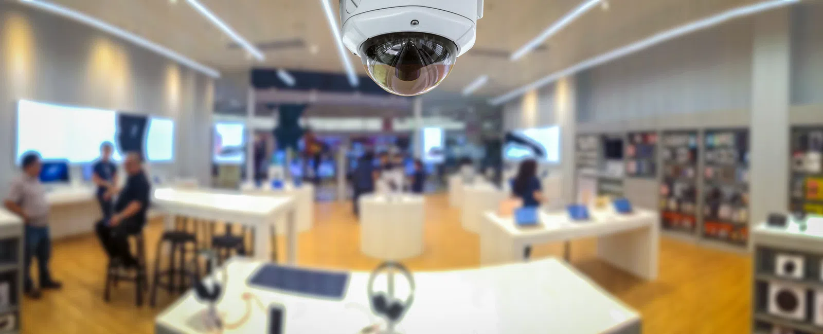 Surveillance camera monitoring bright, modern electronics retail store with customers and digital devices on display.