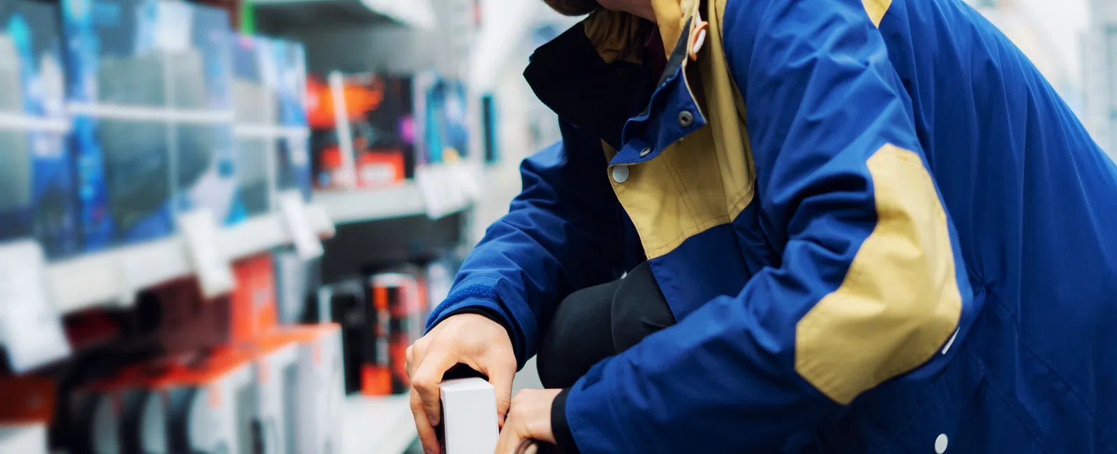 Man in blue and yellow jacket stealing a boxed item from store shelves into his backpack.