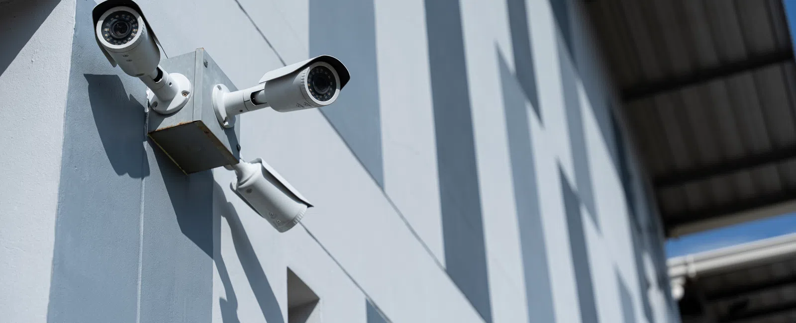 Three white surveillance cameras mounted on a light blue wall under a metal roof casting shadows