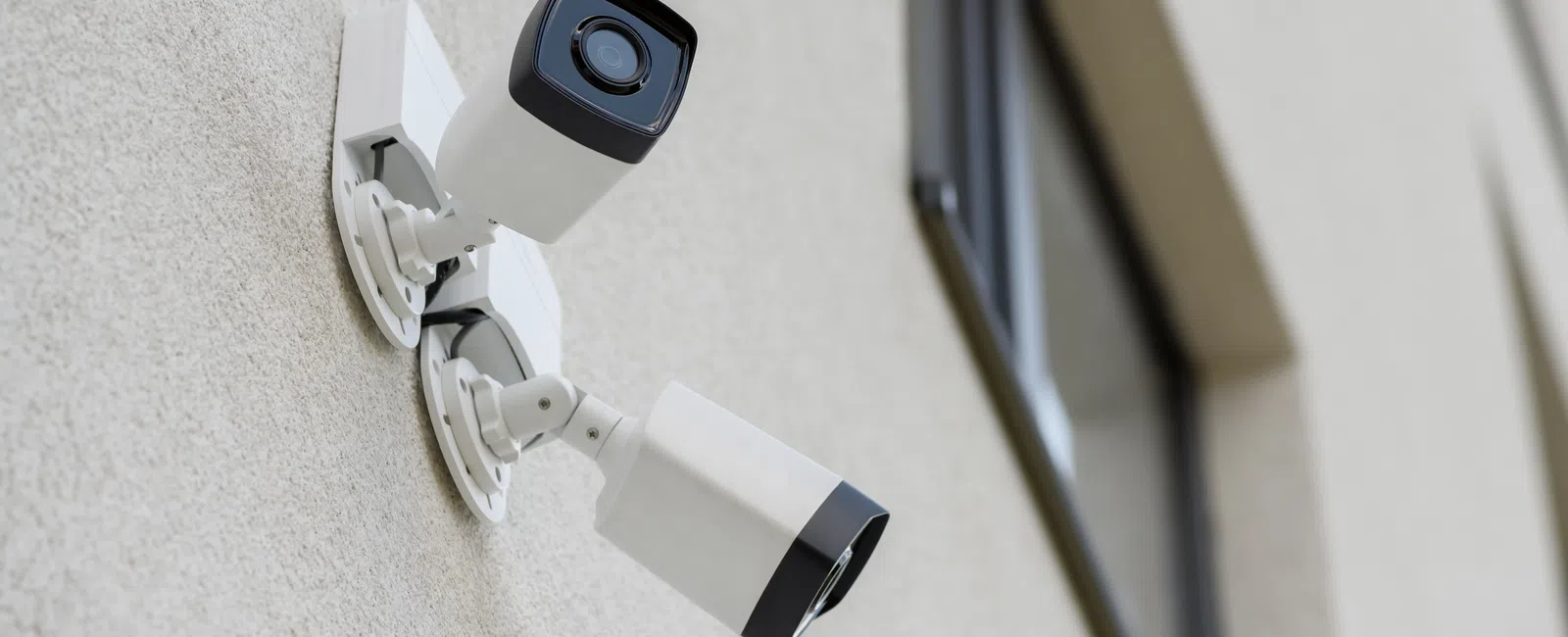 Two white security cameras mounted on a beige building wall near a window for surveillance purposes.