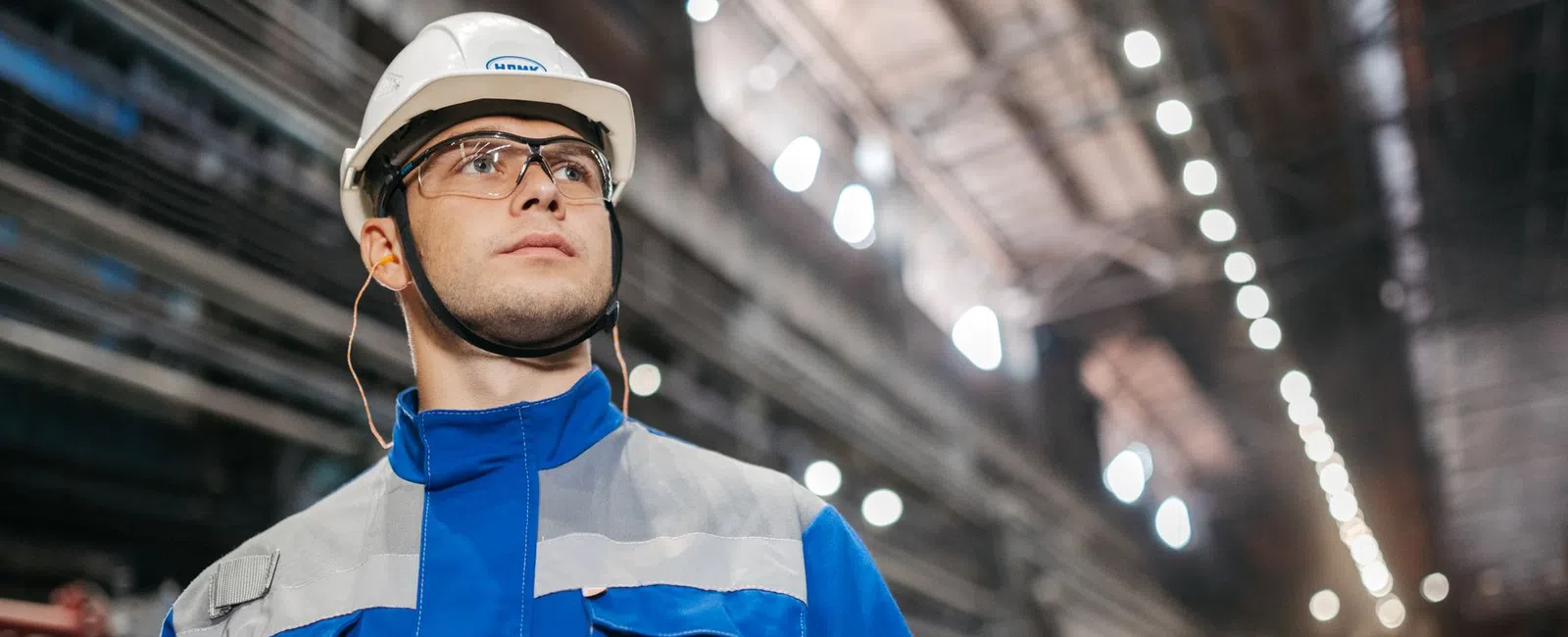 Industrial worker in safety gear holding a tablet inside a large factory or warehouse with bright lights.