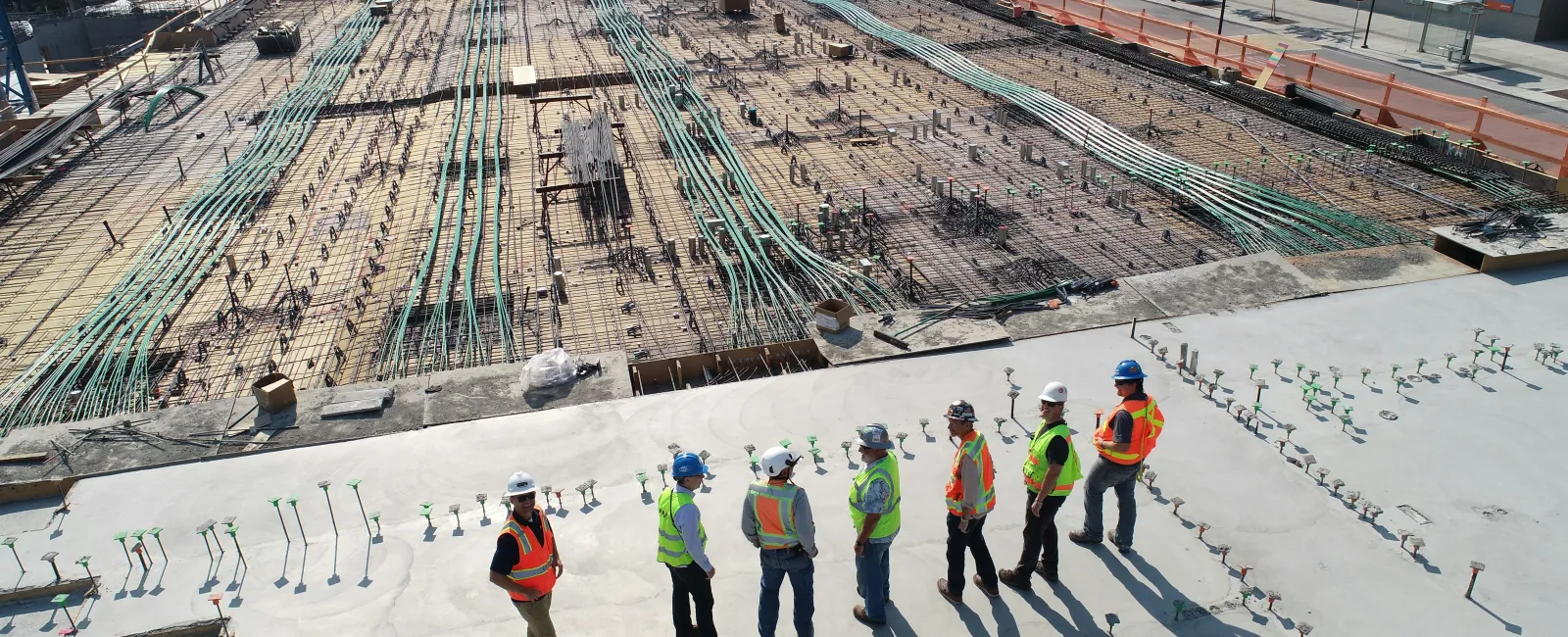 Construction workers in safety gear inspect a large concrete foundation with plumbing and wiring installations on site.