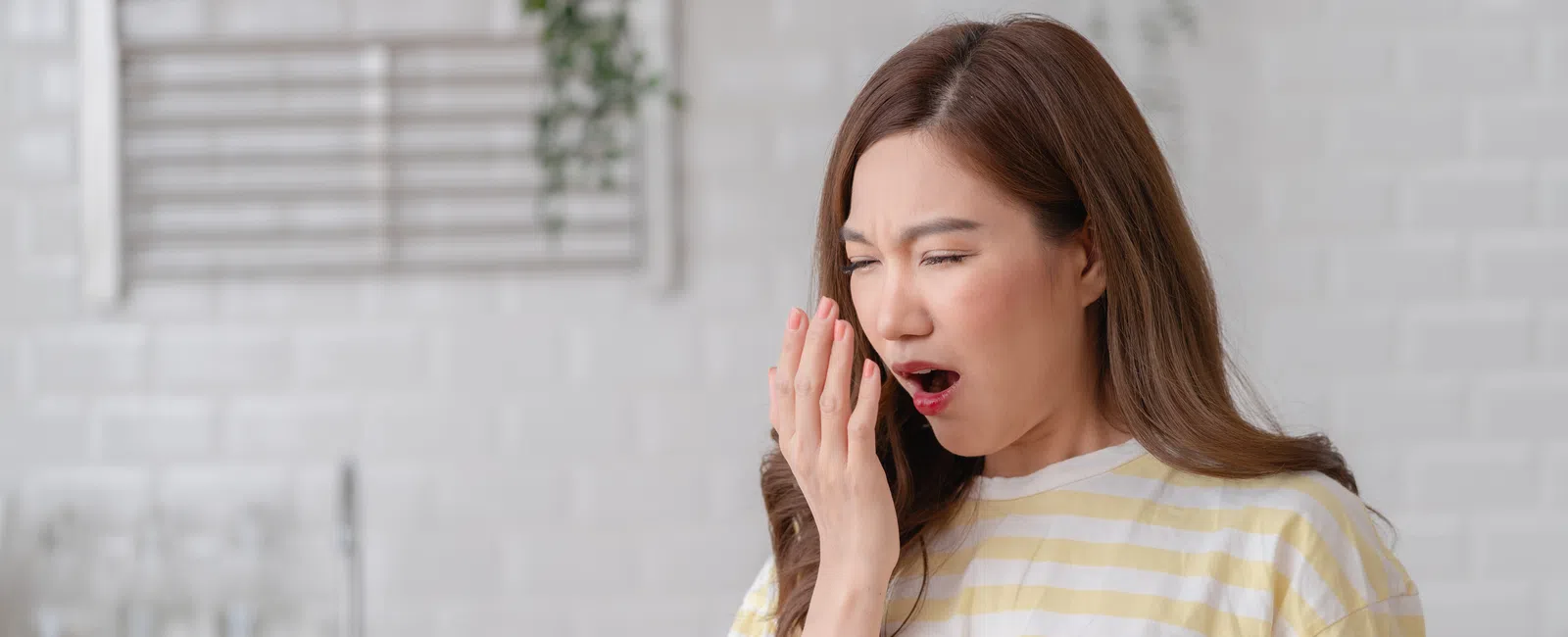Young woman in striped shirt covering her mouth while yawning in a bright kitchen setting