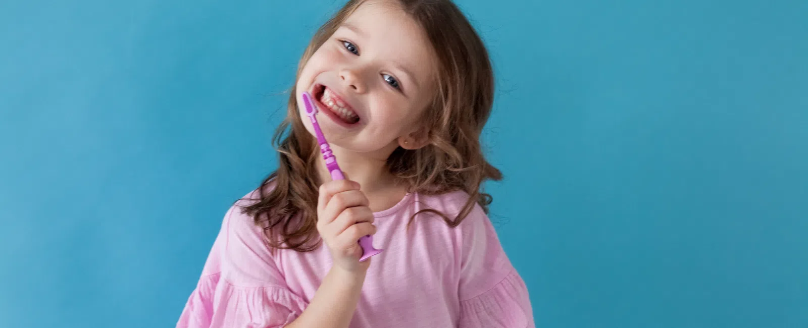 Smiling young girl in pink shirt holding a purple toothbrush against a blue background