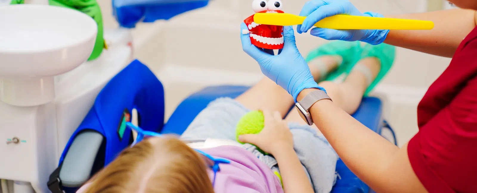 Dentist demonstrates proper brushing on a toy mouth to a child patient lying in the dental chair.