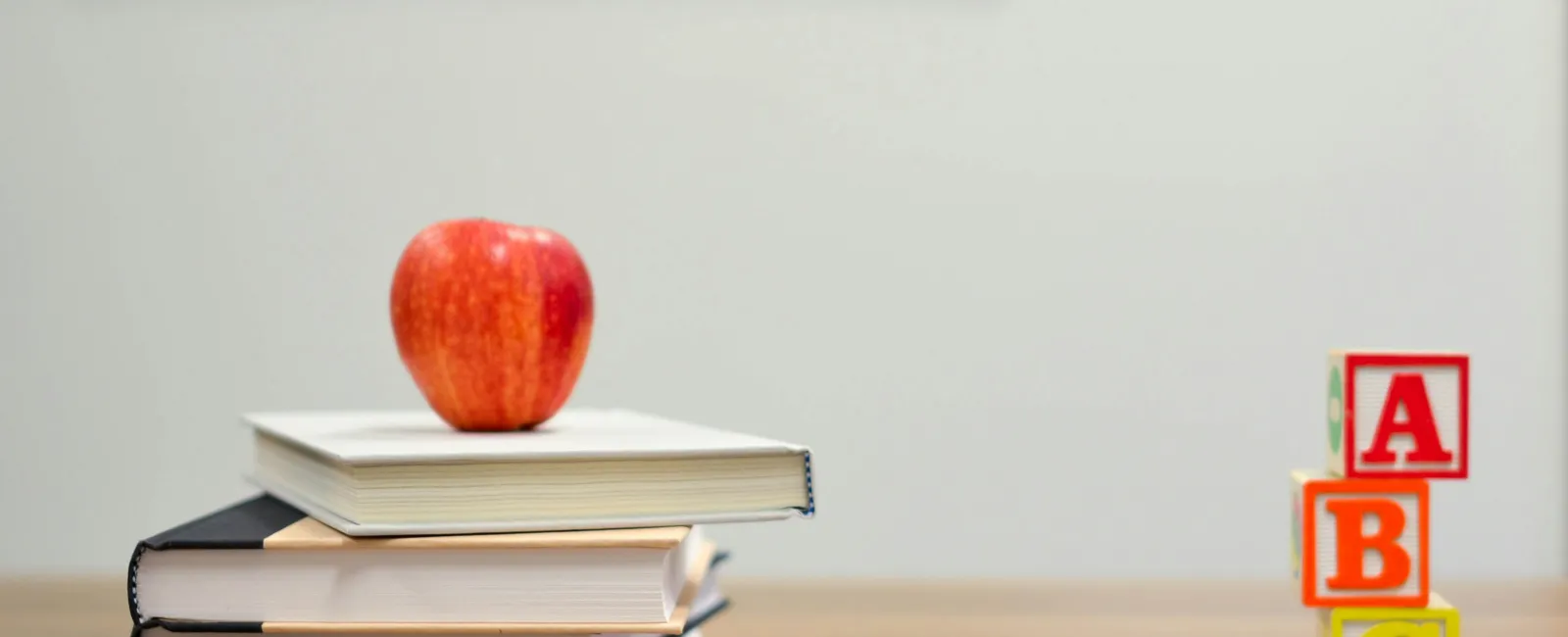 Stack of books with red apple on top, colorful pencils, and ABC blocks on wooden table in classroom setting
