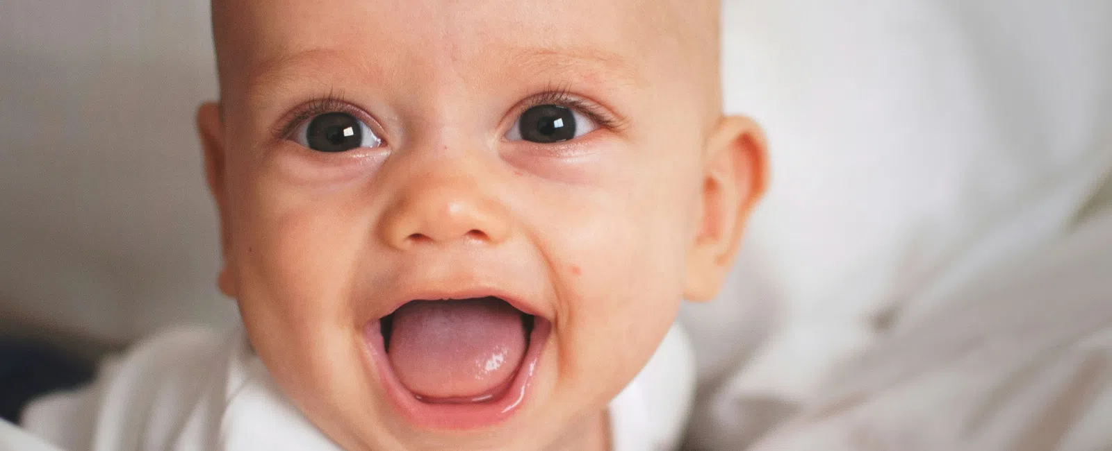 Happy baby with bright eyes and open mouth lying on white bed sheets and smiling joyfully