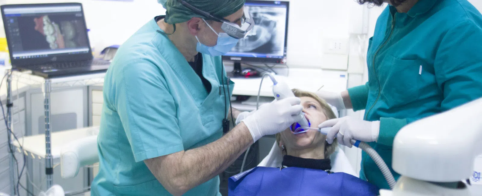 Dentist in scrubs treats patient in dental chair with assistant and digital X-rays on monitors