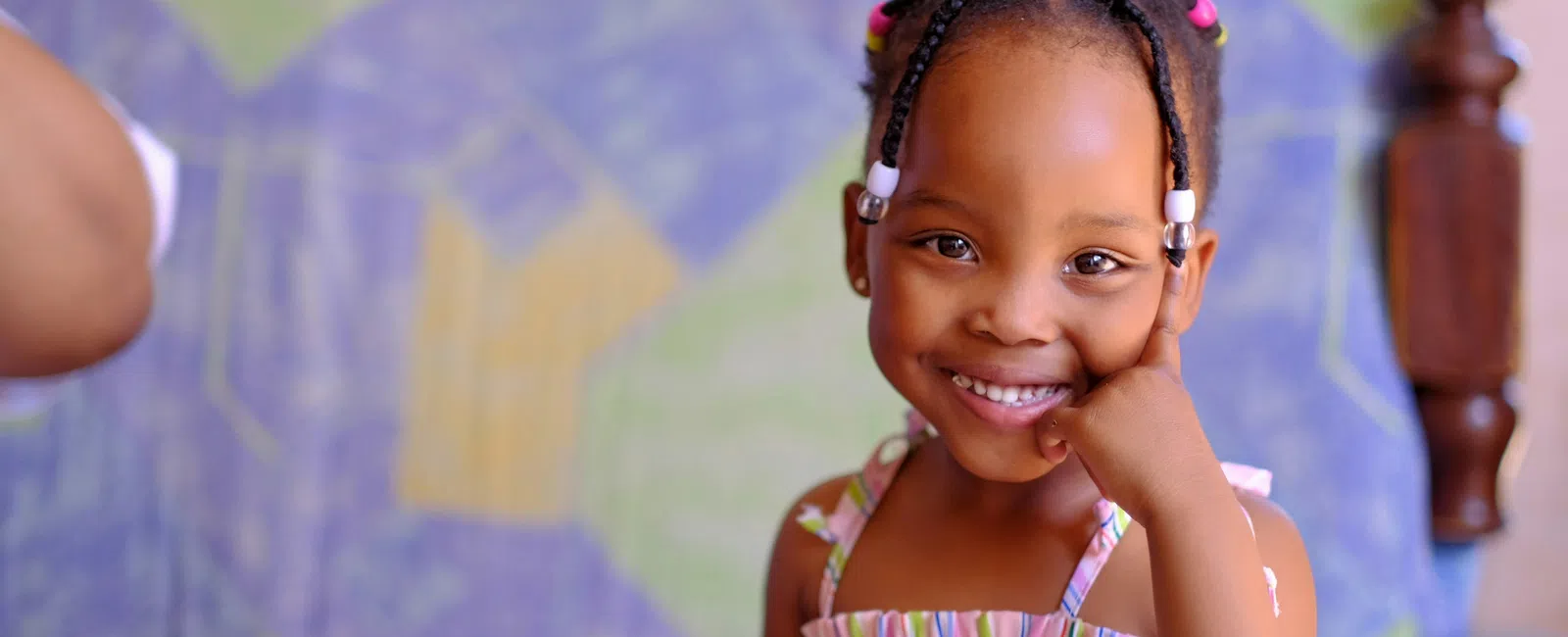 Smiling young girl with braided hair and beads sitting indoors on a patterned sofa smiling at the camera
