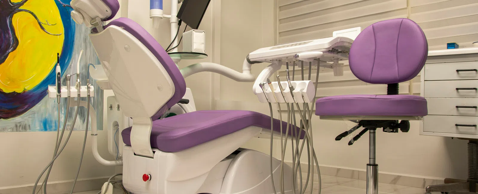 Modern dental chair and stool in purple upholstery inside a clean, well-equipped dental clinic room.