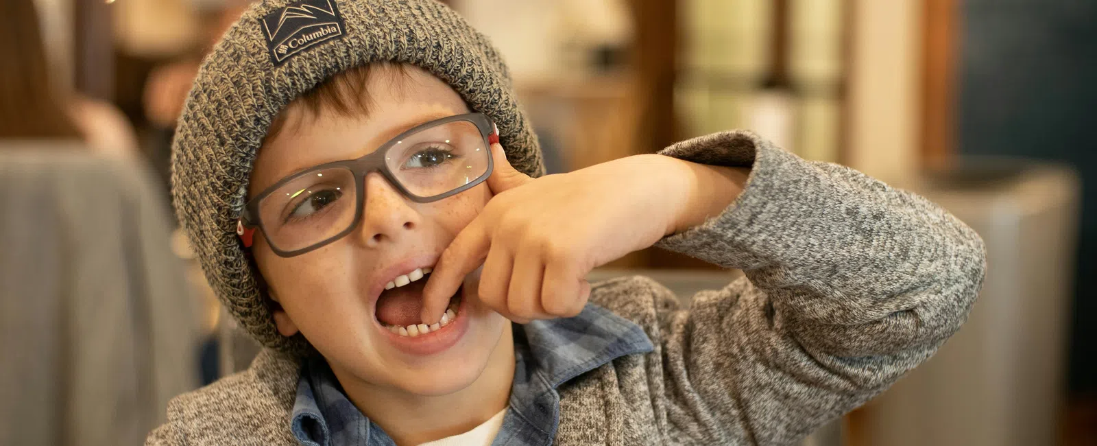 Young boy wearing glasses and knit hat playfully touches his mouth indoors with warm lighting in background