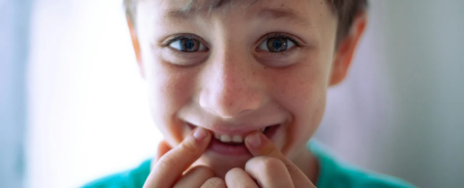 Close-up of a happy young boy with blue eyes and brown hair smiling and pointing at his teeth.