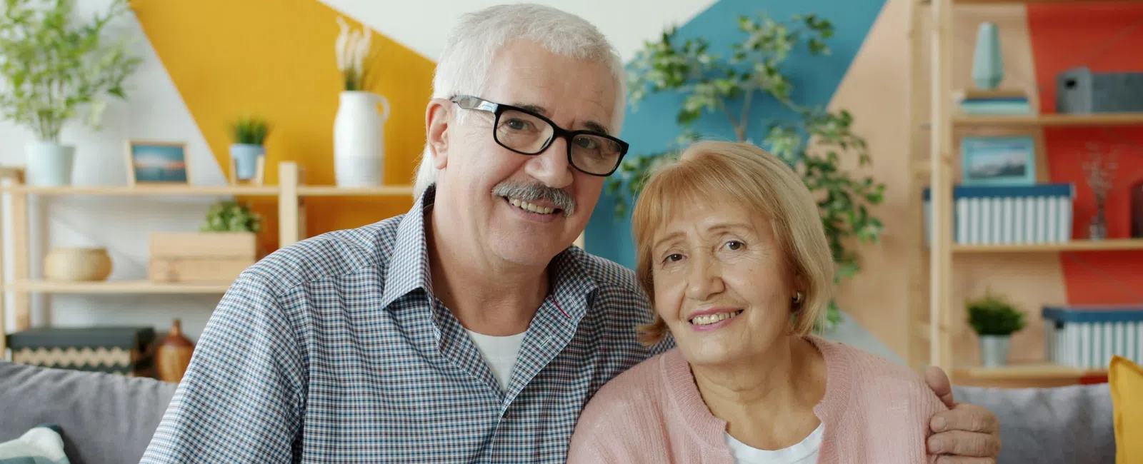 Smiling elderly couple sitting close together on a couch in a colorful modern living room.