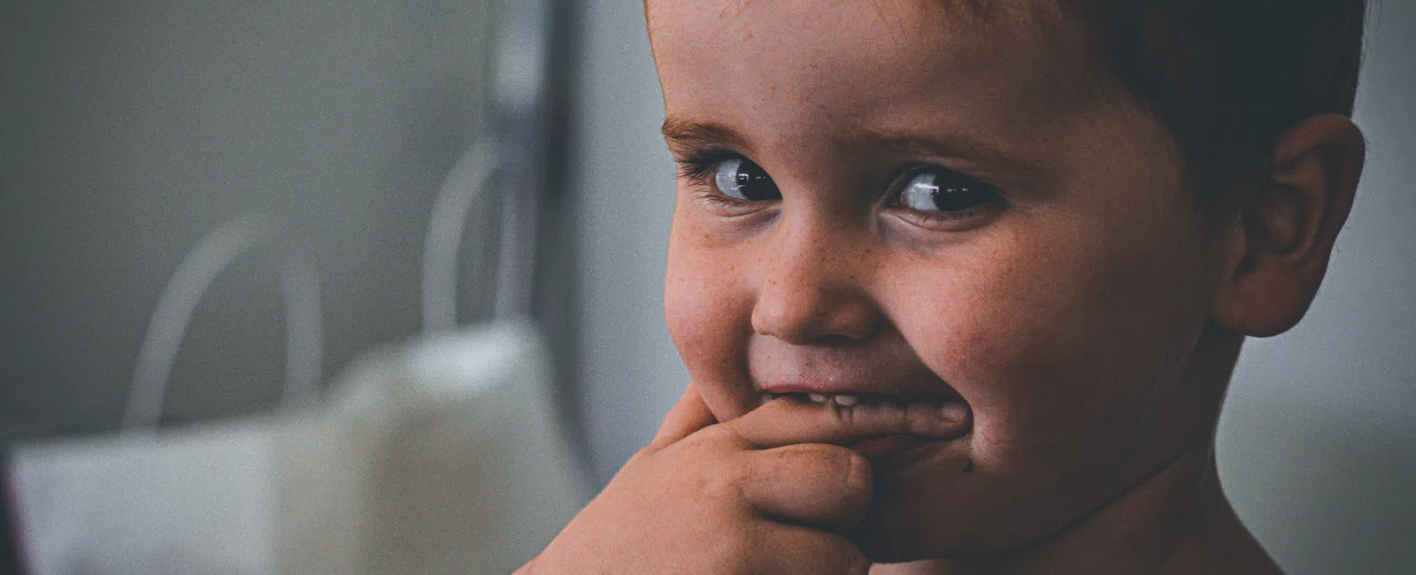 Close-up of a young boy with brown hair smiling playfully with fingers near his mouth indoors.