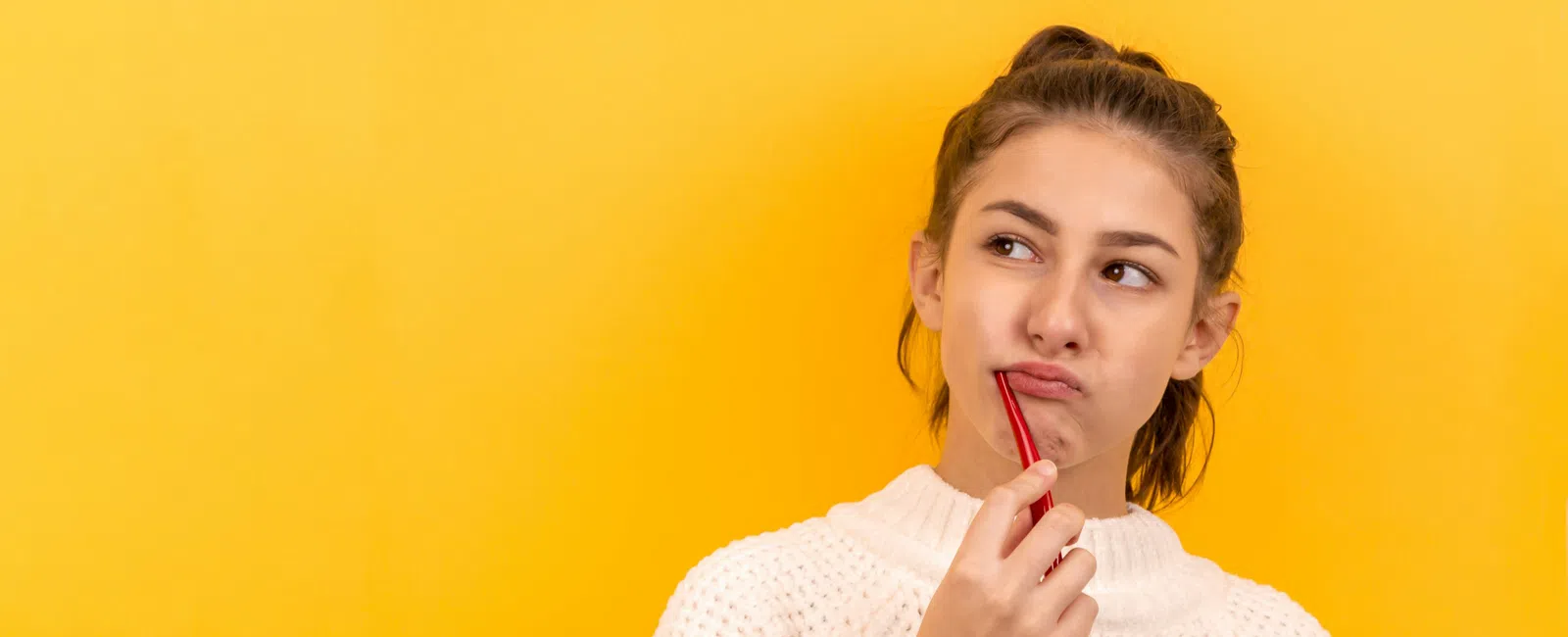 Thoughtful young woman in white sweater holding red pen near lips with bright yellow background.