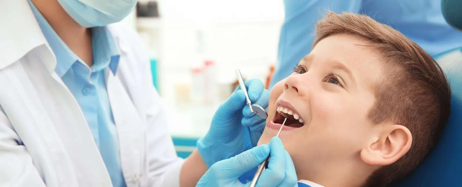 Child smiling while dentist wearing mask and gloves examines his teeth with dental tools.