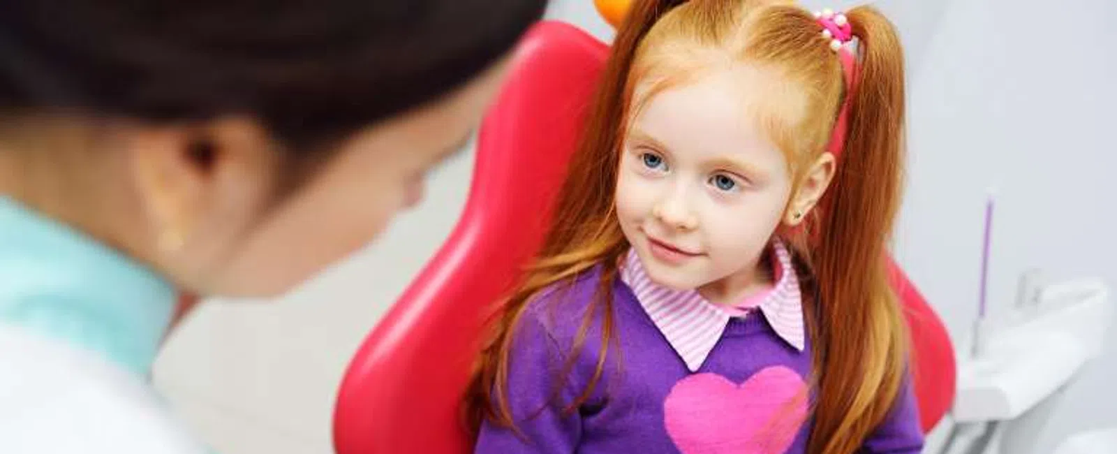 Young girl with red hair learning about dental care from a female dentist holding a mouth model.