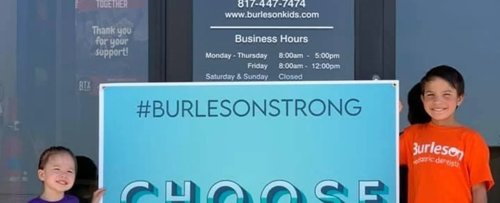 Two children stand beside a blue sign reading Choose kindness outside Burleson pediatric dentistry office.