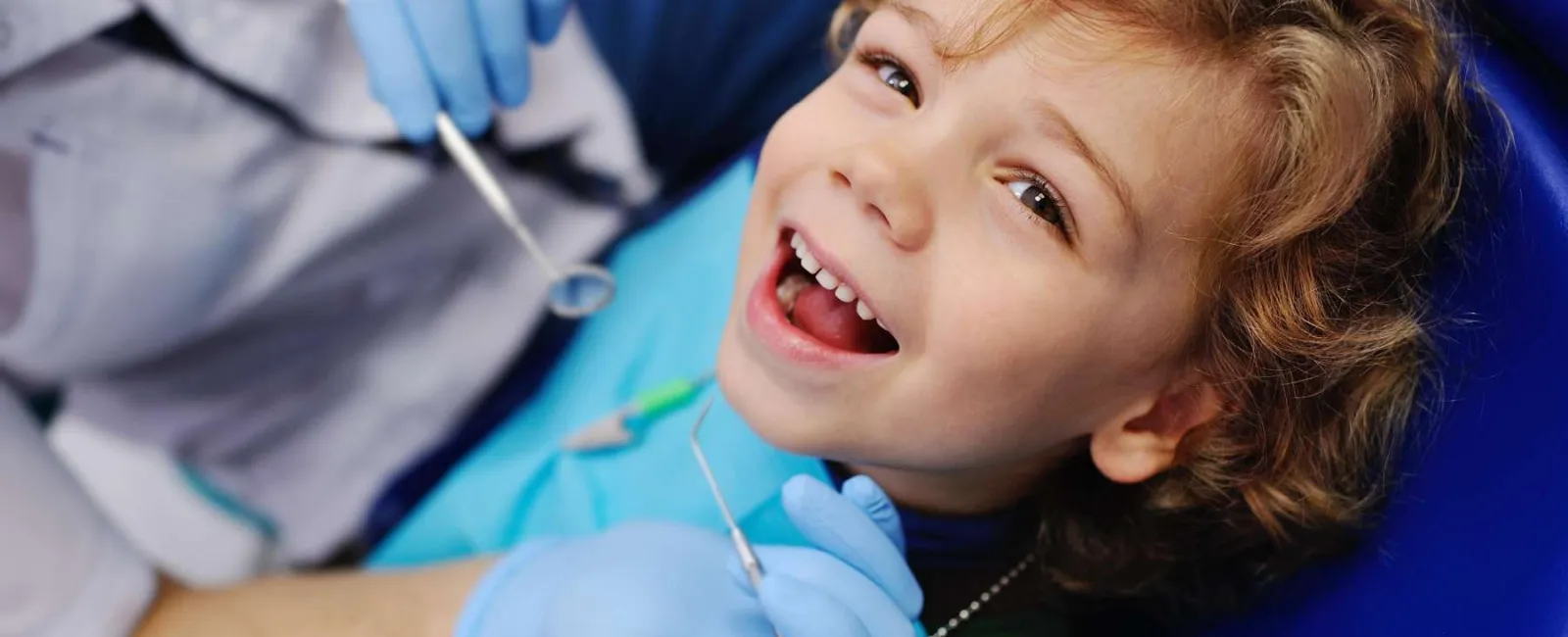 Smiling child with open mouth receiving dental checkup from dentist wearing blue gloves.
