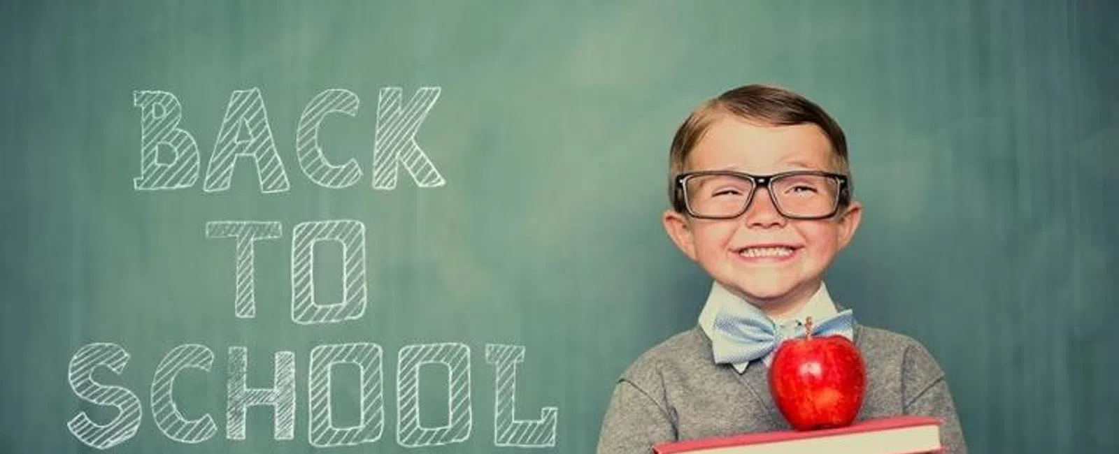 Smiling young boy with glasses holding books and an apple, standing in front of a chalkboard with Back to School text.